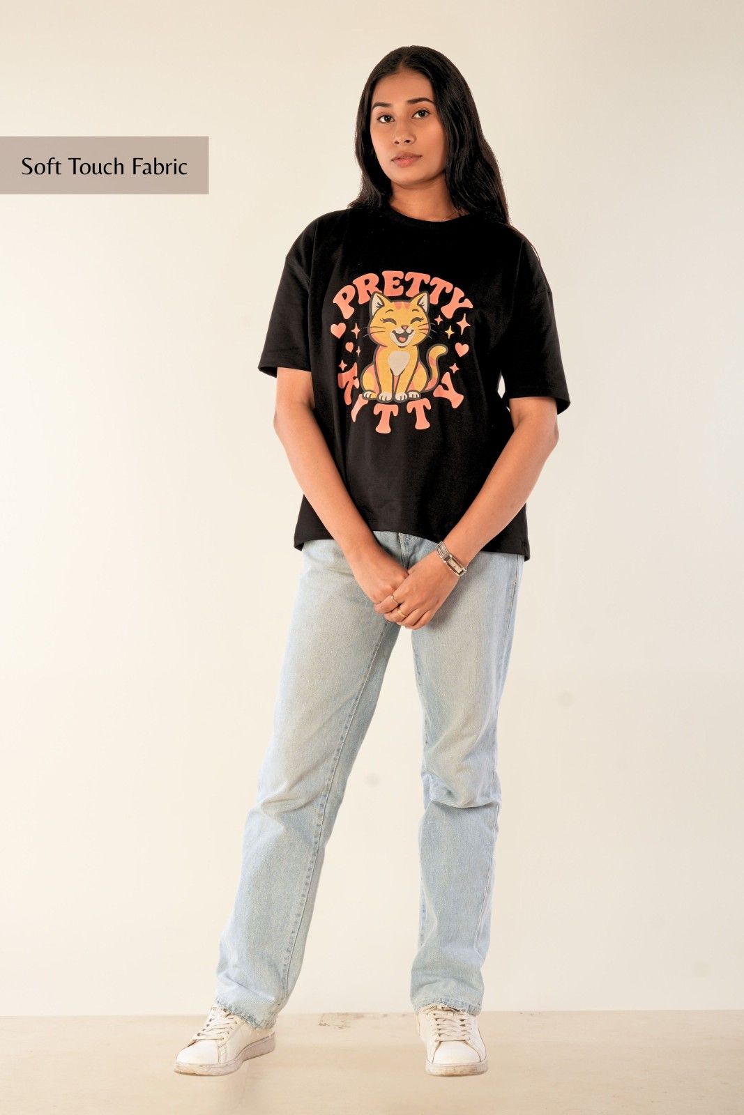 Black oversized women's t-shirt with Pretty Kitty graphic design. Cute cat illustration in peach tones with hearts and stars. Premium screen-printed streetwear from Berrys & Cigaredes. Full body styling with light blue denim and sneakers.
