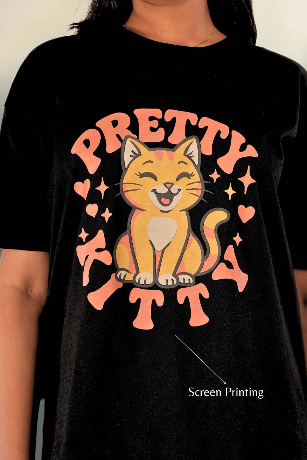 Chest close-up of black oversized women's t-shirt featuring Pretty Kitty cat graphic. Screen-printed design with cute cat illustration in peach tones, hearts, and stars. Premium print detail from Berrys & Cigaredes casual streetwear collection.
